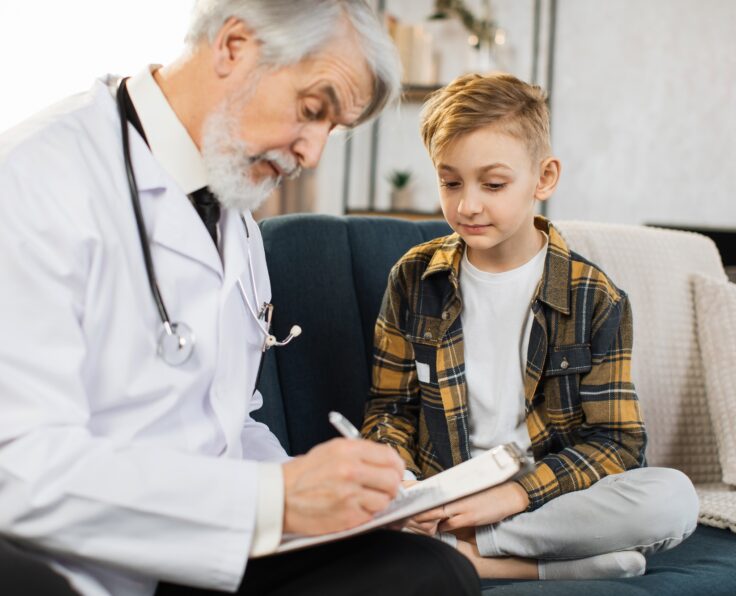 Caring mature man doctor, consulting cute sick boy, making notes in clipboard